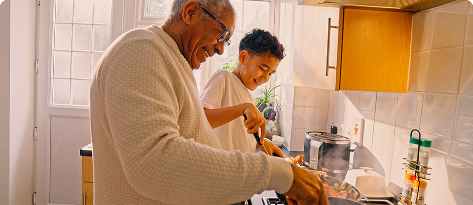 An older man and a young boy smiling as they cook together at a stovetop in a sunlit home kitchen, stirring food in a pan with steam rising.