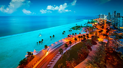 Aerial panoramic view of a tropical beachfront city with turquoise waters, palm-lined boulevard, and glowing city lights at dusk