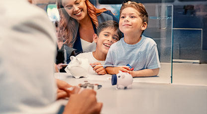 Image of a woman and two children with piggy banks at a bank counter exchanging coins with a banker