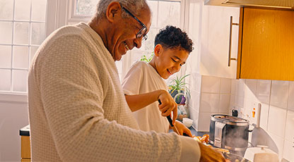 An older man and a young boy smiling as they cook together at a stovetop in a sunlit home kitchen, stirring food in a pan with steam rising.