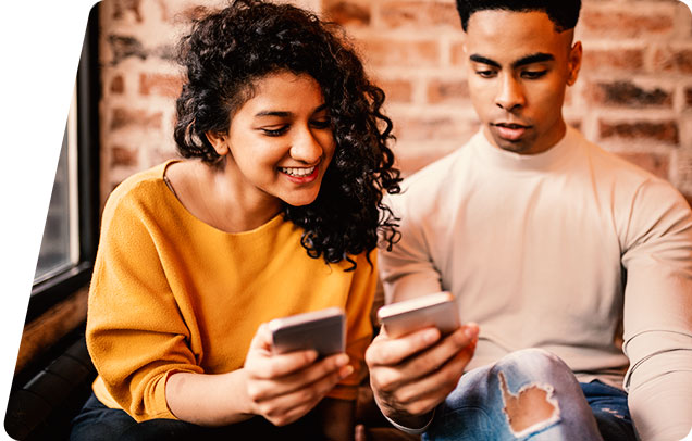 Two young people looking at each other's smartphone in front of an exposed brick wall