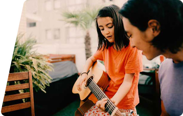 A young person plays the guitar while their older sibling smiles and listens