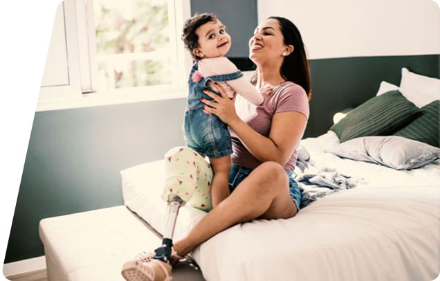 "A smiling woman with a prosthetic leg sits on a bed, joyfully holding up a laughing toddler in a denim outfit.