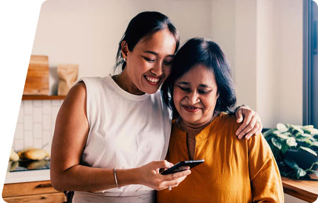 A younger adult has their arm around an older adult as they look at something on their smartphone