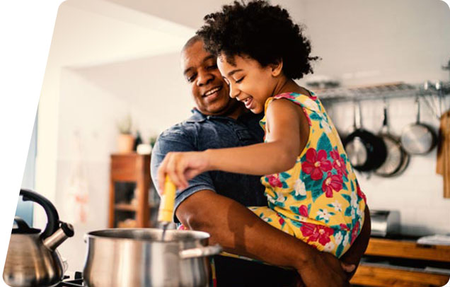 A parent holds a smiling child stirring something in a pot on the stove while the other parent watches