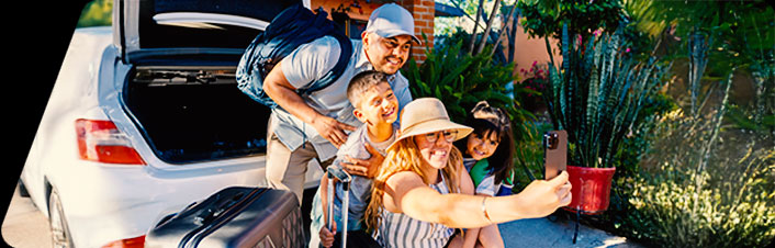 A happy family taking a selfie together beside the open trunk of a white car packed with luggage, surrounded by lush greenery on a sunny day.