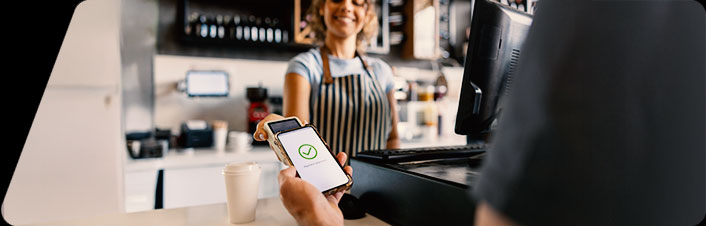 A customer holding a smartphone displaying a green checkmark, completing a contactless mobile payment at a café counter.