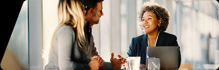 "A professional woman smiling and speaking with two colleagues during a meeting at a bright office table with a laptop nearby.