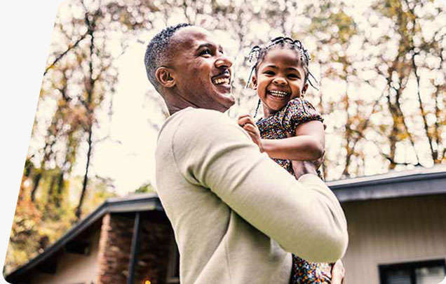 An adult holds a smiling child outside in front of a house on an autumn day.