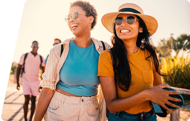 Two young women smiling and walking together outdoors on a sunny day, both wearing sunglasses, with other people visible in the background.
